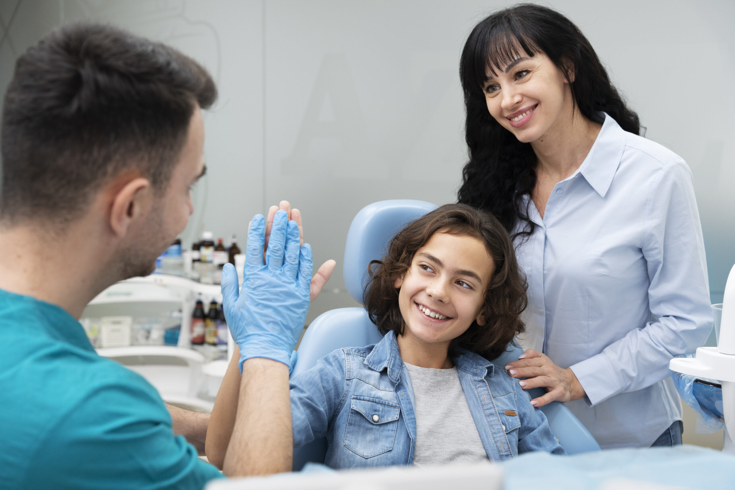 Photo d'illustration de la catégorie Pour les plus jeunes du thème "nos conseils" avec un enfant aux cheveux mi-longs installés dans le fauteuil de dentiste, sa mère à ses côtés, checkant la main gantée du dentiste. Tous sont souriants